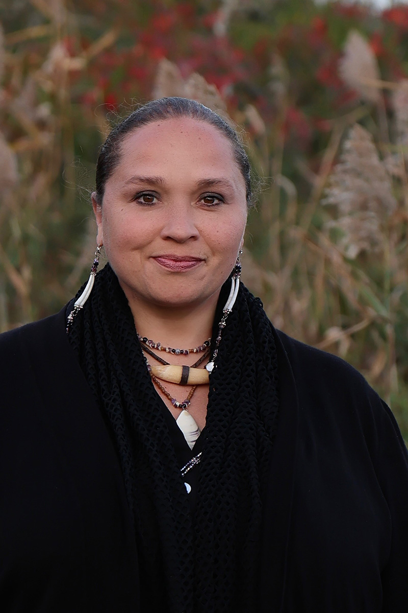 Courtney M. Leonard, a woman, in a black outfit stands outdoors on Shinnecock Nation territory, surrounded by tall grass. She wears indigenous jewelry and has a peaceful expression, with a backdrop of greenery.