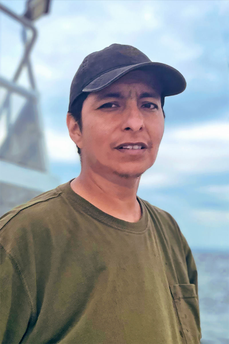 A chest-up portrait of Vick Quezada, a brown-skinned man in a brown baseball cap and olive green t-shirt, smiling and looking slightly off-camera against a blurred backdrop of a boat and misty water. Photo by Jamie Tam.