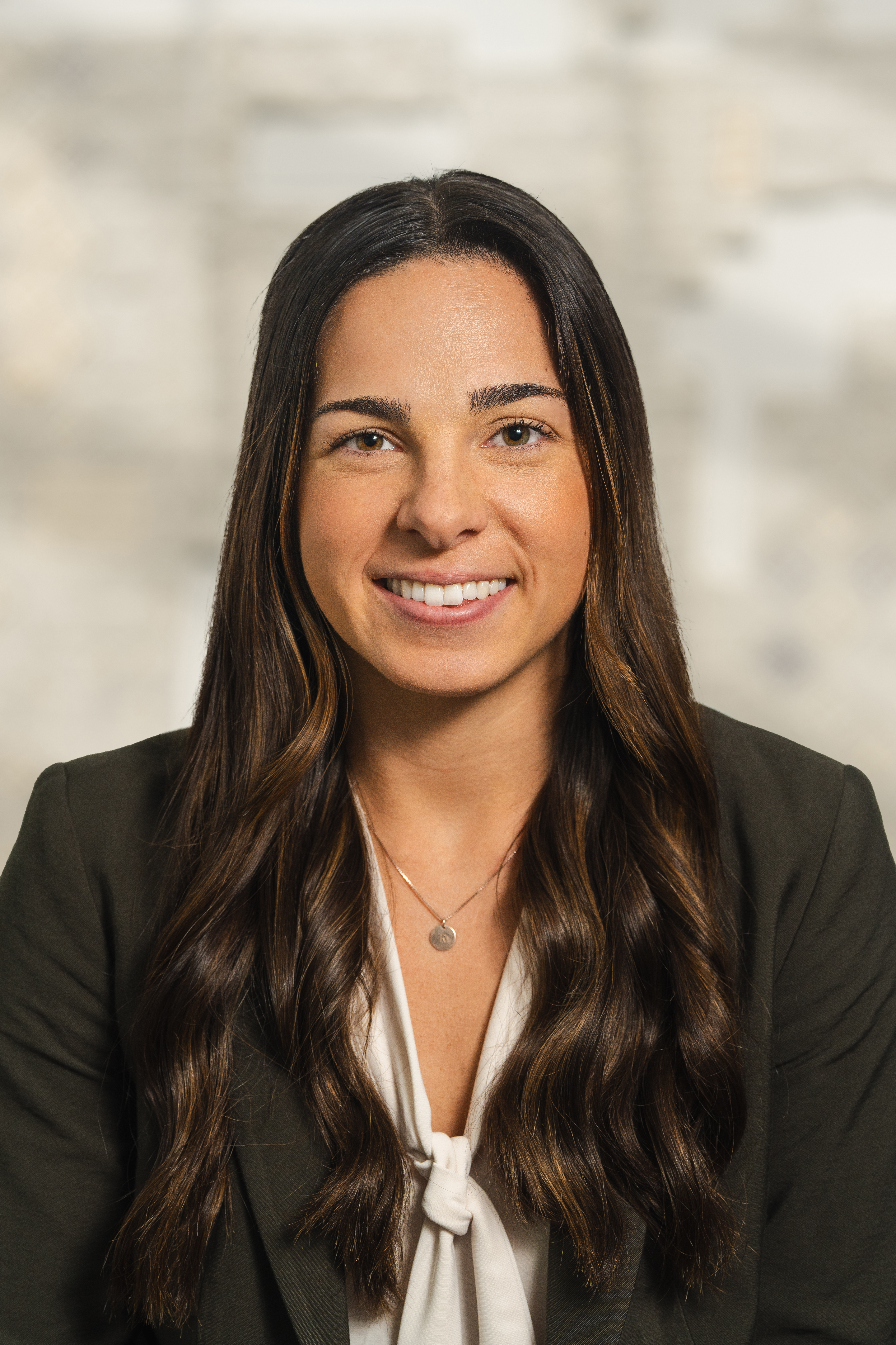 Brianna Goodfellow, with long dark brown curly hair, smiling while standing against a grey backround 