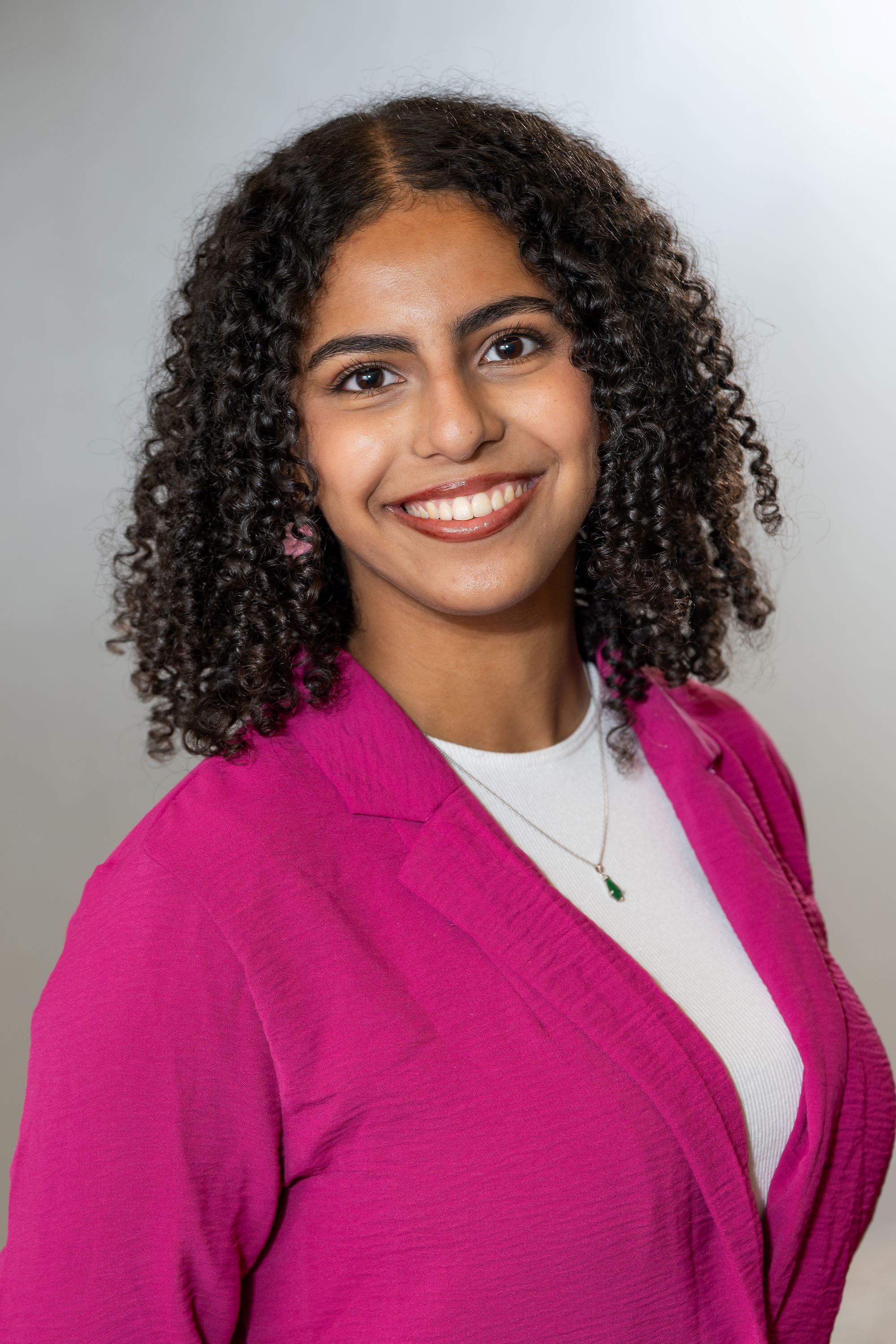 Portrait of a smiling person wearing a bright pink blazer, with curly hair and a pendant necklace.