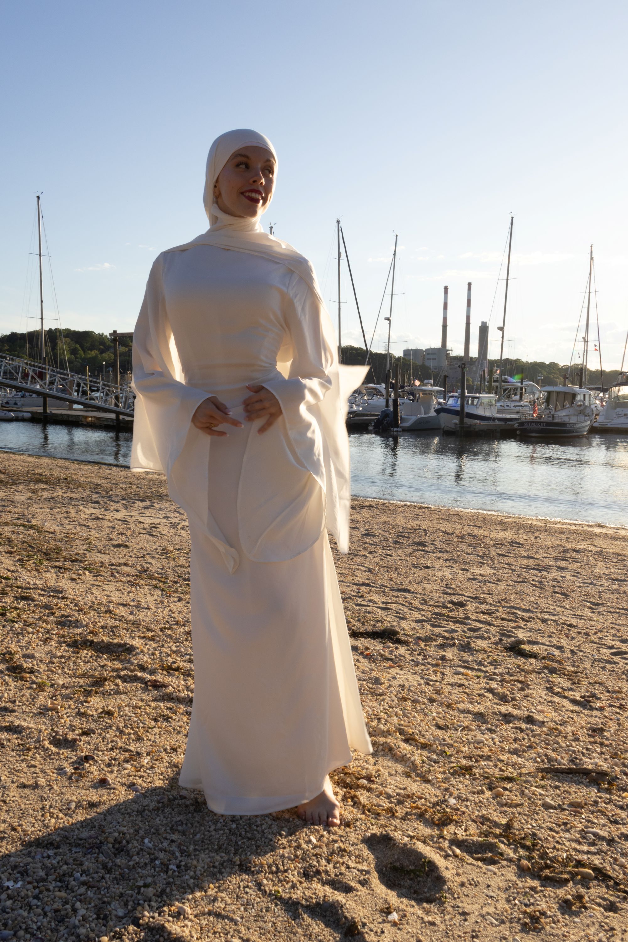 A woman in a flowing white dress and headscarf stands barefoot on a sandy beach. She is smiling, with sailboats and calm water behind her at sunset.