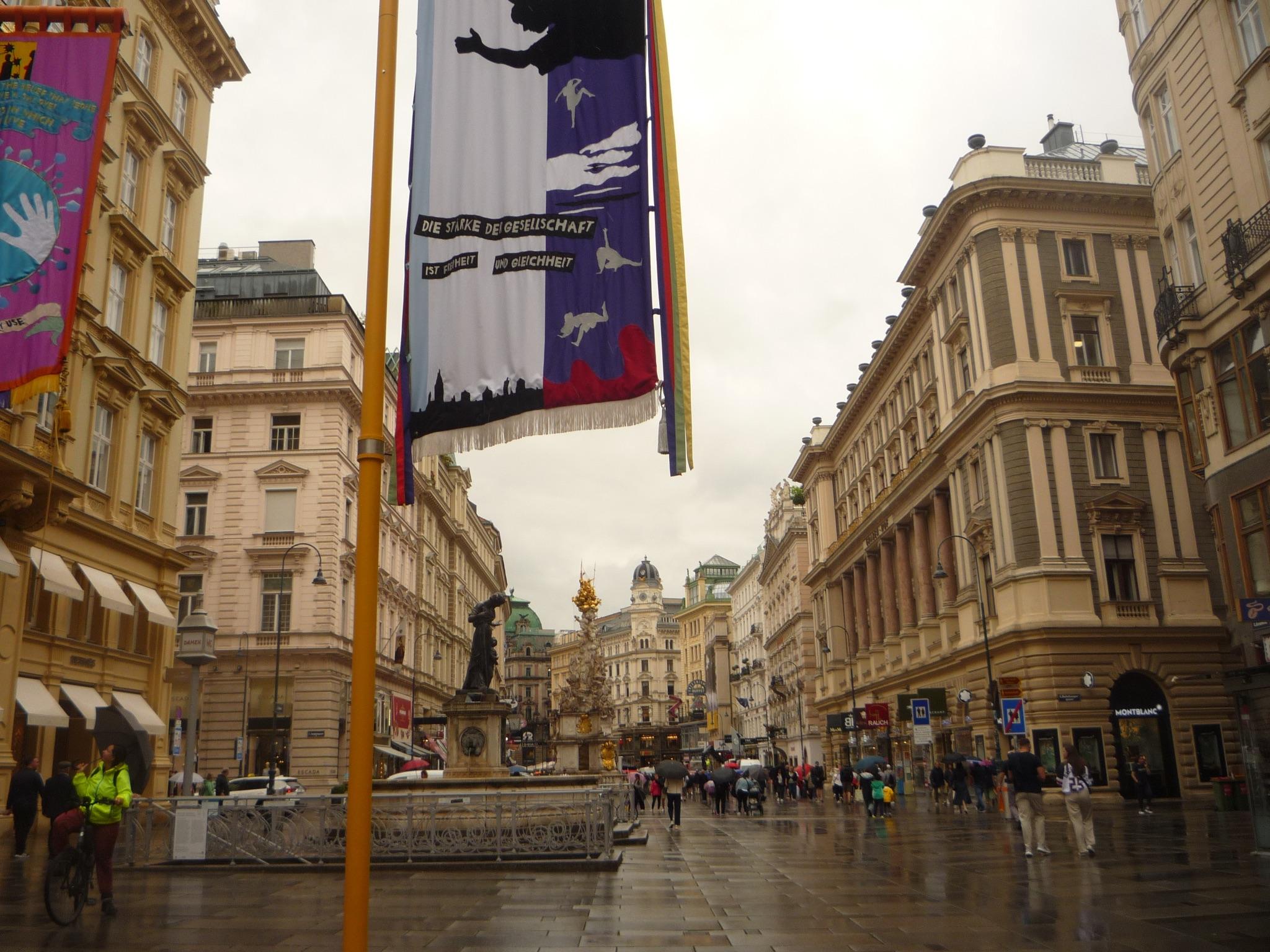 View of a busy pedestrian street in Vienna, lined with neoclassical buildings and decorated with colorful banners, under a cloudy sky.