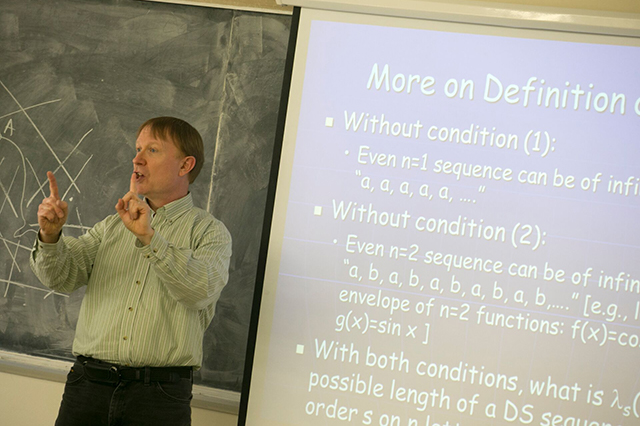Applied Mathematics and Statistics Faculty A man lectures in a classroom, gesturing towards a projected slide with mathematical text. A chalkboard with diagrams is in the background. Educational atmosphere.