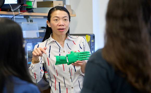 Biomedical Engineering Faculty A person in a patterned shirt holds up a green prosthetic hand, engaging in discussion with two others. The setting is a laboratory, conveying learning and innovation.