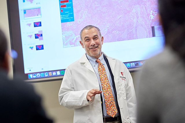 Biomedical Informatics Faculty A man in a white lab coat presents confidently in front of a screen displaying scientific data and images, conveying an educational tone.