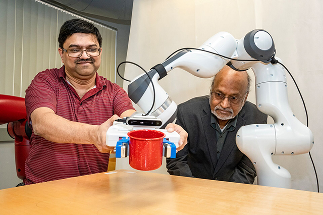 Computer Science Faculty Two men interact with a robotic arm gripping a red mug on a table. One man smiles while the other observes, showcasing technology and human interaction.