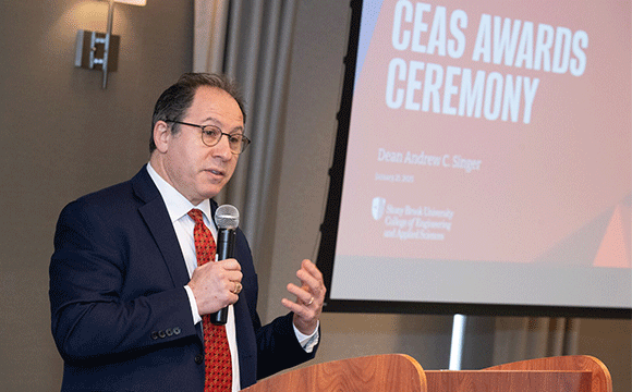 A man in a suit speaks at a podium with a microphone, gesturing with one hand. A presentation slide titled "CEAS Ceremony" is visible in the background.