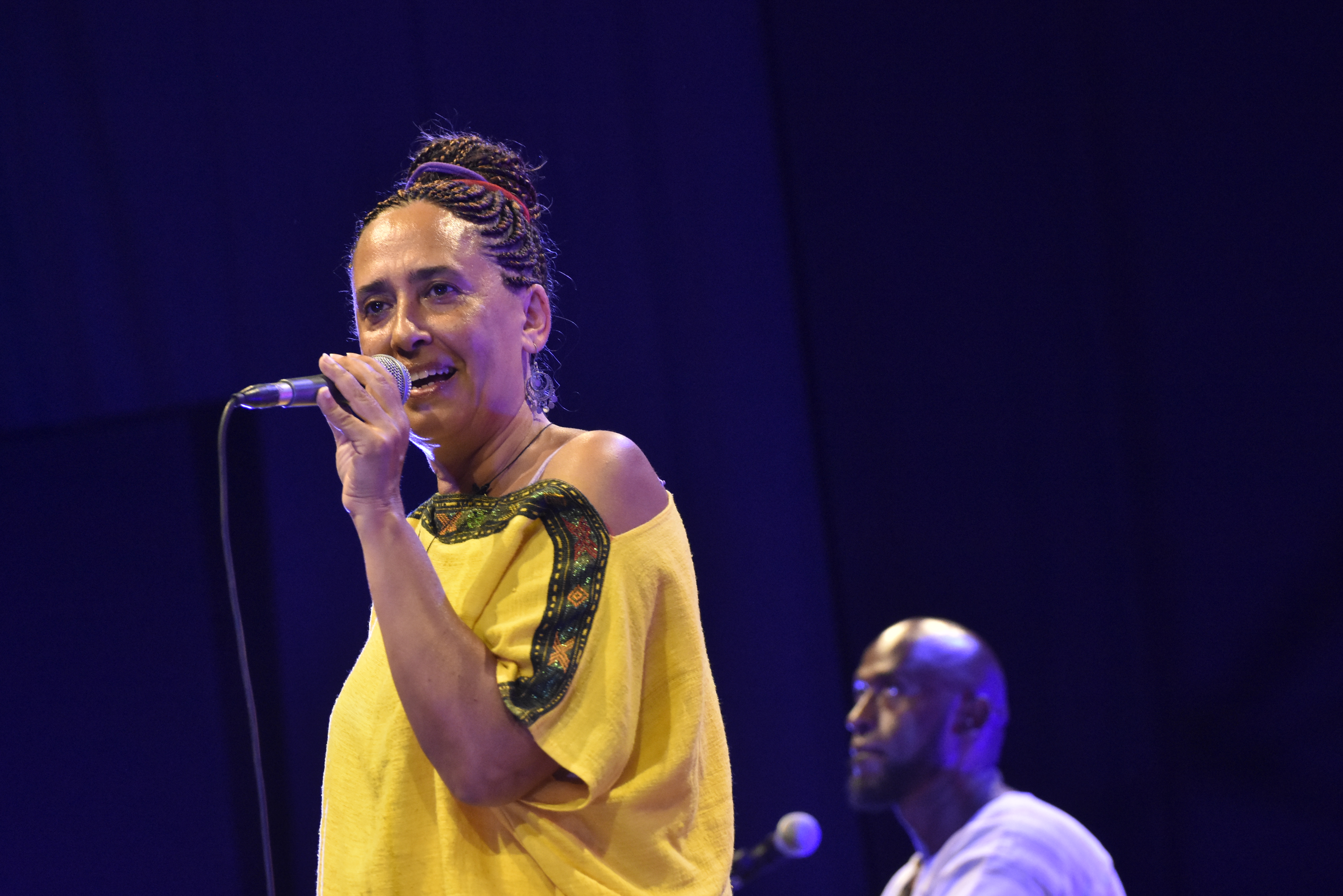 snapshot of singer, performer, and writer Gabriella Ghermandi in a live performance. She is holding a microphone and singing and a man behind her is playing an instrument