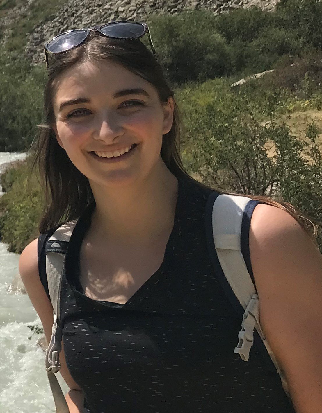 A young woman with long brown hair and a bright smile stands outdoors in a natural setting. She is wearing a dark, speckled tank top and a white backpack. Her sunglasses are pushed up onto her forehead. In the background, a rushing mountain stream and rocky, brush-covered terrain are visible under bright sunlight.