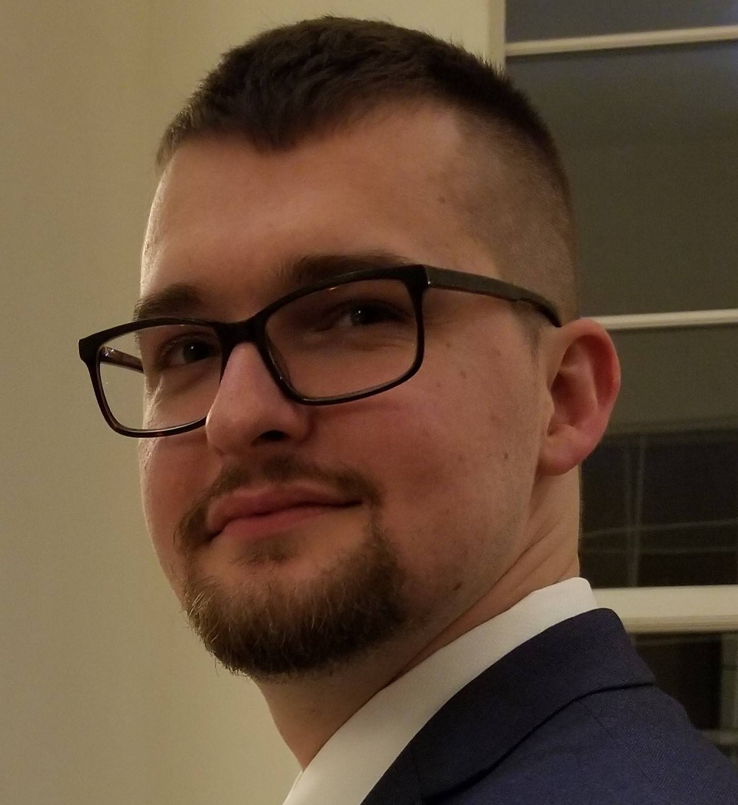 Asberry_Headshot A close-up, chest-up headshot of a young man with light skin, short-cropped dark hair, and a neatly trimmed goatee. He is wearing dark-rimmed rectangular glasses and is angled slightly to his right, looking toward the camera with a subtle smile. He is dressed professionally in a white collared shirt and a dark blue suit jacket. The background is a simple, neutral-toned wall with a hint of a window frame or architectural detail to the right.